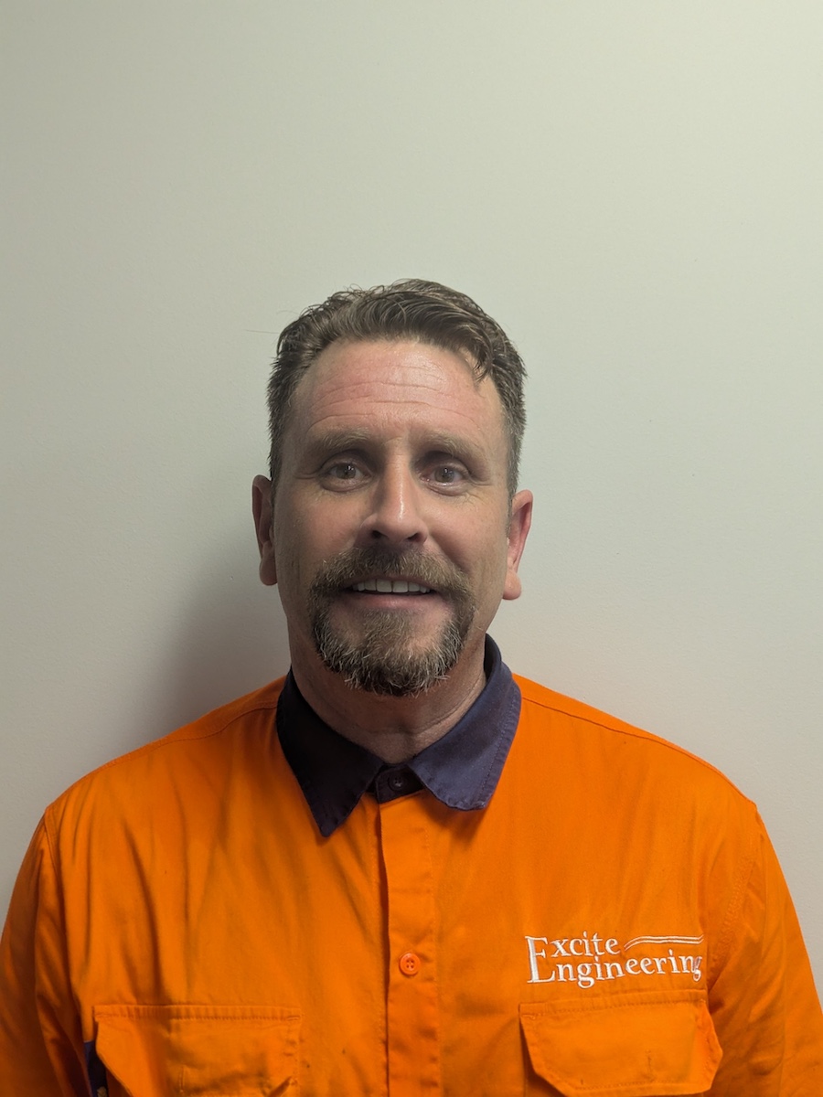A man with short brown hair and a goatee smiles at the camera, wearing an orange shirt with 