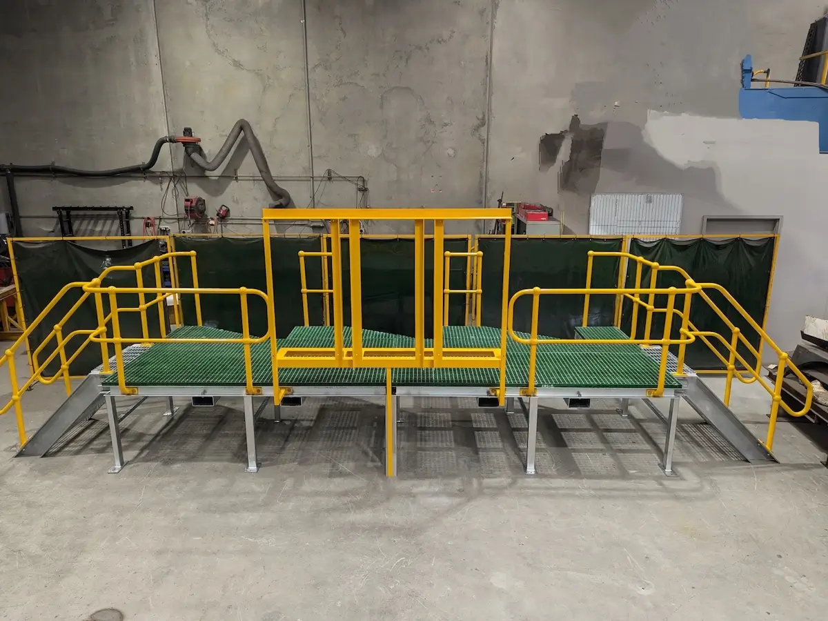 A metal work platform with yellow railings and green grated steps, featuring two staircases on each side, sits in an industrial warehouse with concrete floors and walls.