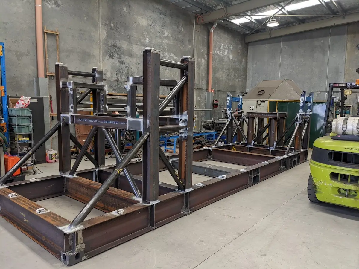 Large metal frame structure with welded steel beams and supports inside an industrial workshop. Various tools, equipment, and a green forklift are visible in the background.