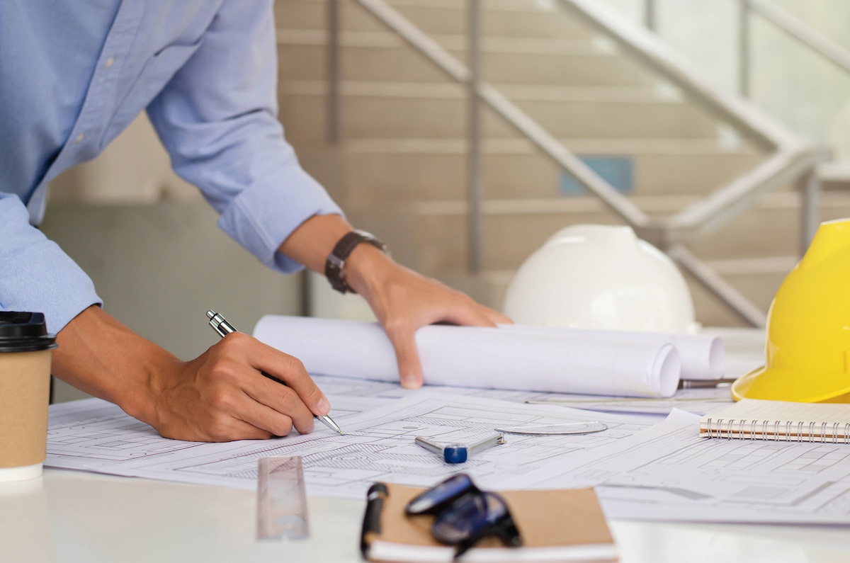 A person in a blue shirt is drawing on architectural blueprints at a desk, surrounded by rolled plans, a coffee cup, safety helmets, a ruler, eyeglasses, and a notepad, with stairs visible in the background.