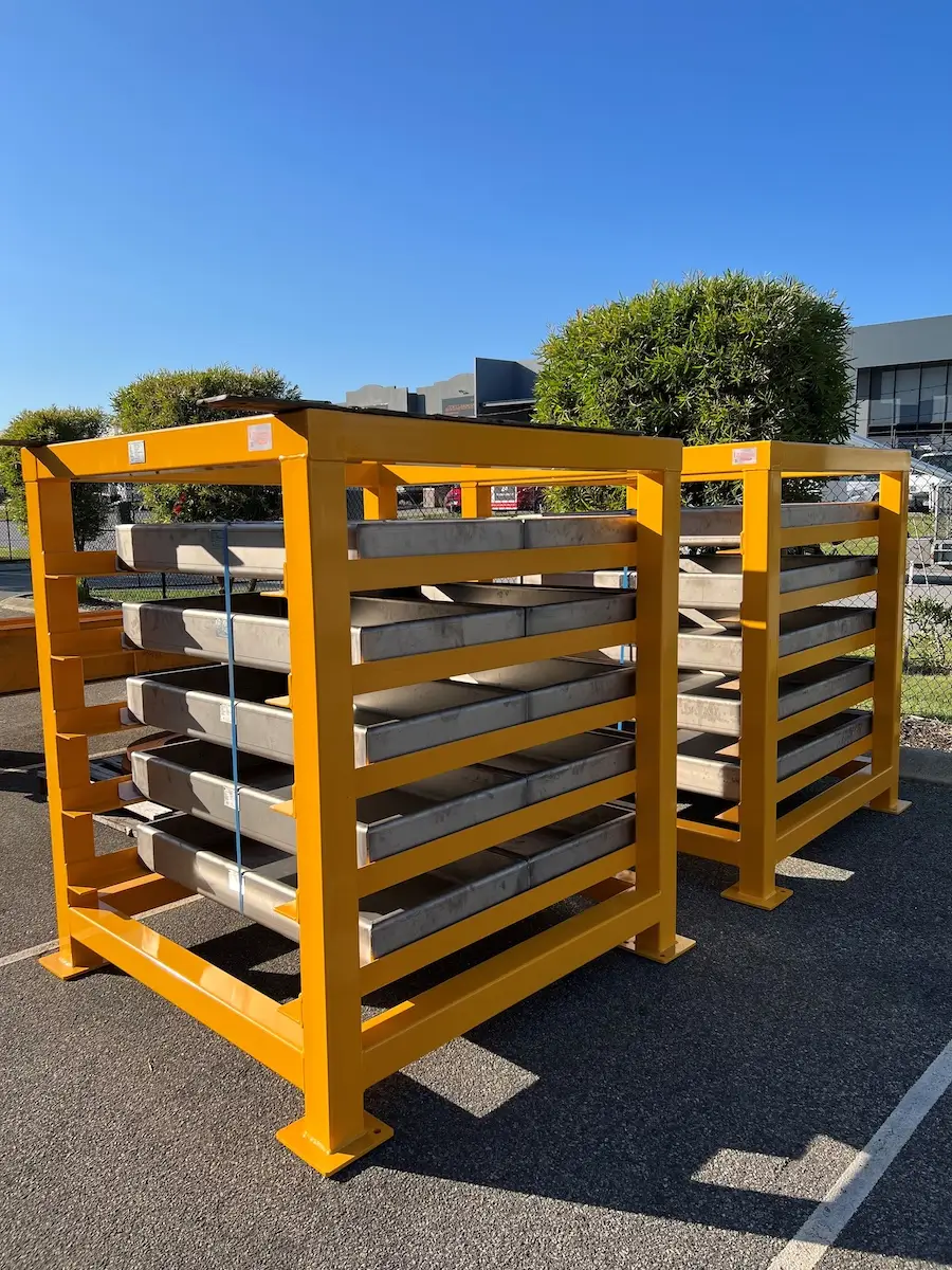 Two large yellow metal storage racks hold stacked rectangular metal sheets, secured with straps, outdoors in a parking lot under a clear blue sky. Trees and industrial buildings are visible in the background.