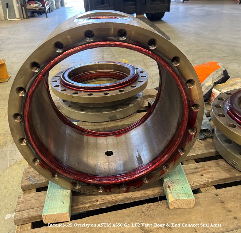 A large metallic valve body with a visible red Inconel-625 overlay lining the internal seal area, resting on wooden blocks in an industrial workshop setting. Other valve components are visible in the background.