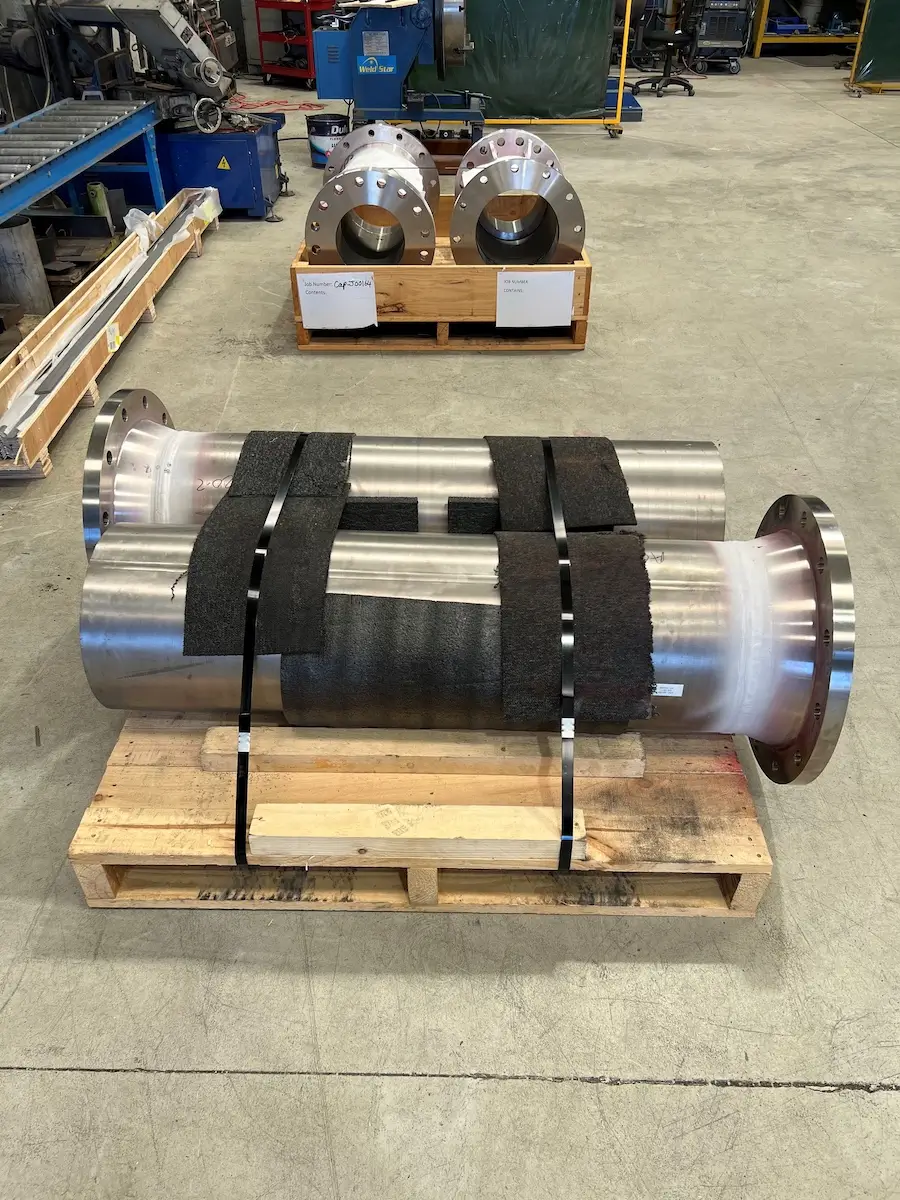 Two large metal pipes with flanges are strapped to a wooden pallet with black bands and padding. In the background, two similar flanged pipes are placed on another pallet in an industrial workshop setting.