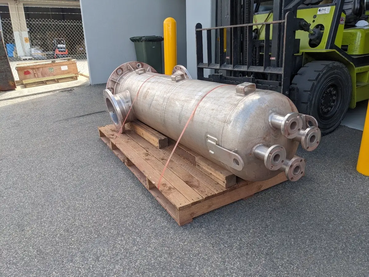 A large, cylindrical metal pressure vessel with several nozzles lies secured with straps on a wooden pallet beside a forklift in an outdoor industrial area.