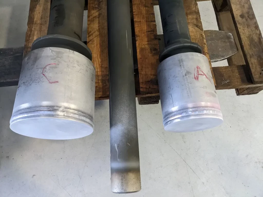 Two large metal pistons labeled "C" and "A" rest on a wooden pallet, with a dark cylindrical rod placed between them on a light-colored floor.