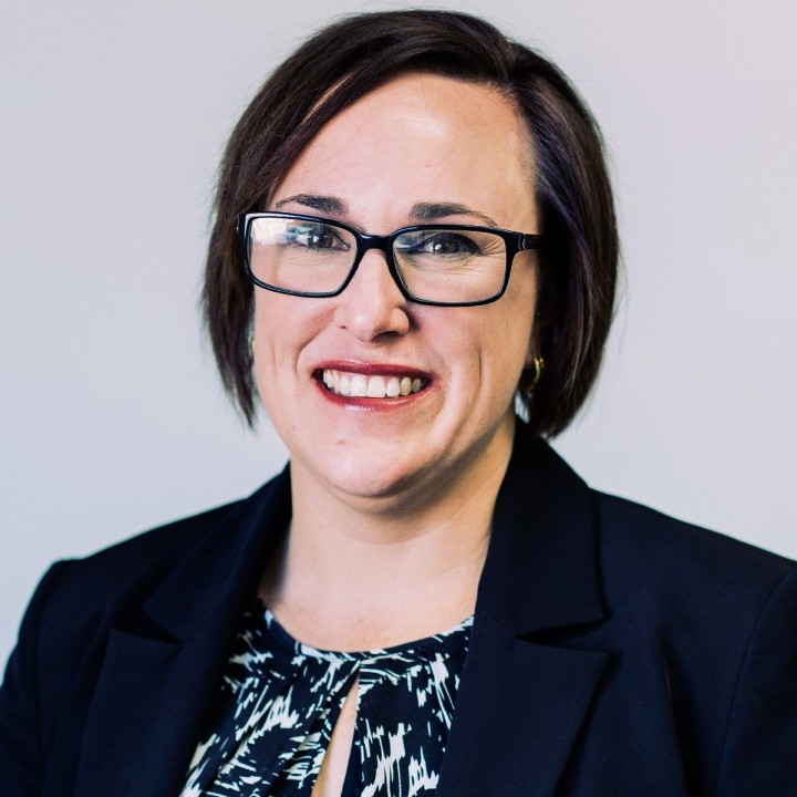 A woman with short brown hair and glasses smiles at the camera. She is wearing a dark blazer over a patterned blouse and standing in front of a light background.