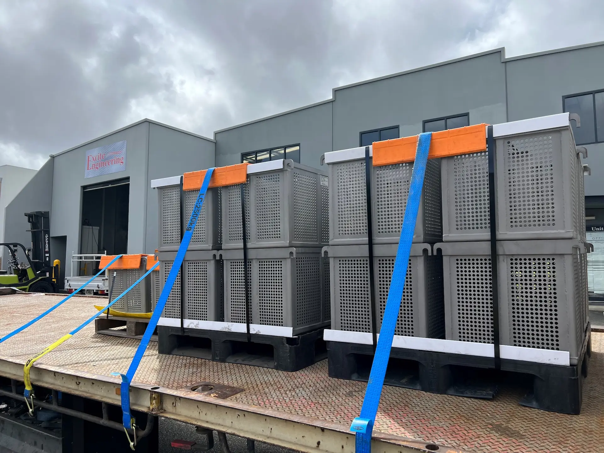Large gray crates secured with blue and yellow straps are loaded on a flatbed truck outside a modern industrial building under a cloudy sky.