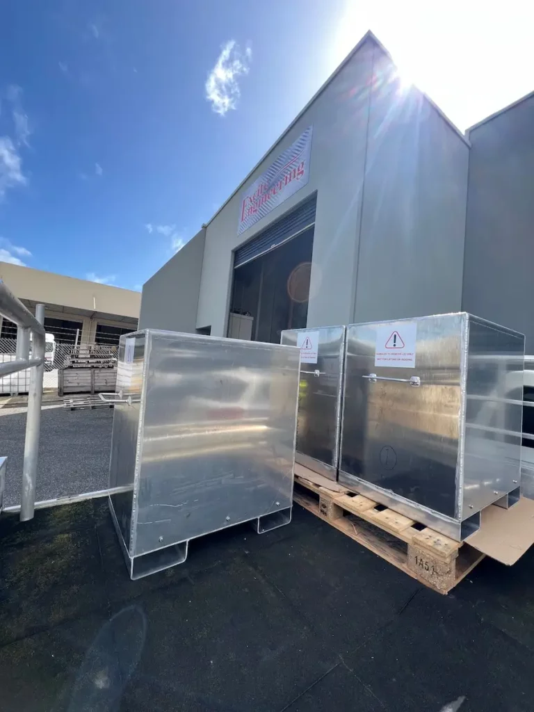 Two large metal containers sit outside a gray industrial building under a blue sky with clouds. The containers are on a pallet, and warning signs are visible on their sides. The building has a loading dock and signage above.