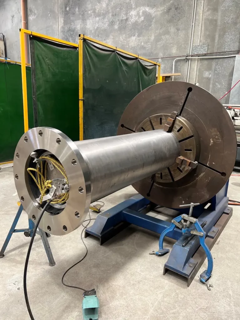 A large cylindrical metal machine part is mounted horizontally on a blue stand in an industrial workshop, with cables connected to one end and a green safety screen in the background.
