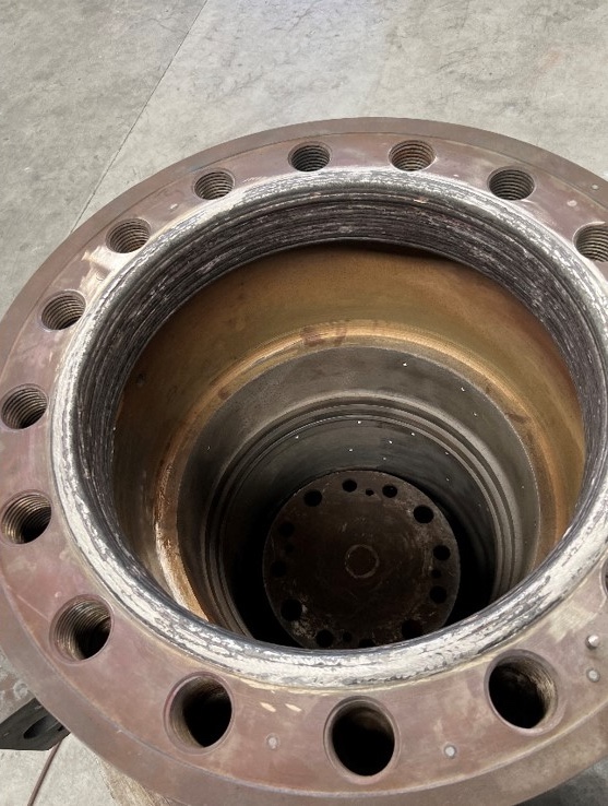 A close-up view of a large, cylindrical metal component with a flanged rim and multiple bolt holes, showing signs of wear and a textured inner surface, set on a concrete floor.