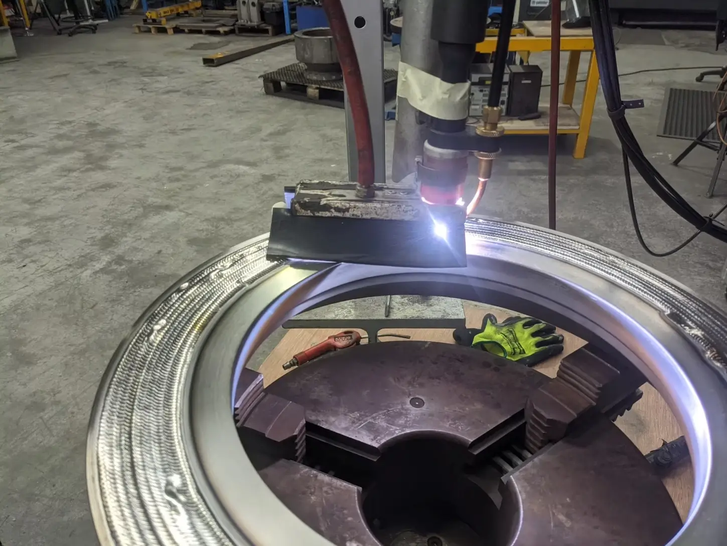 A welding torch is working on a large circular metal piece in an industrial workshop. A pair of gloves, a small wrench, and other tools are visible nearby on the workbench.